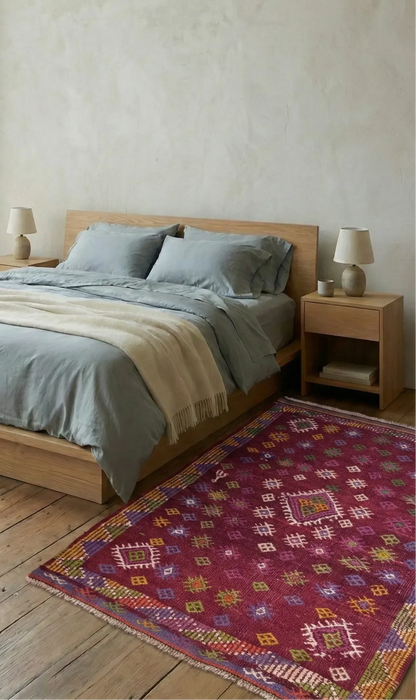 A vibrant magenta 40 years old hand woven flatwoven 100% wool natural dye Turkish rug with colorful geometric motifs and a central diamond medallion, styled on aged wood floors in the Master Bedroom of the Amsterdam Loft Studio, featuring a wooden platform bed and sheer curtains in a Scandi-Boho aesthetic.